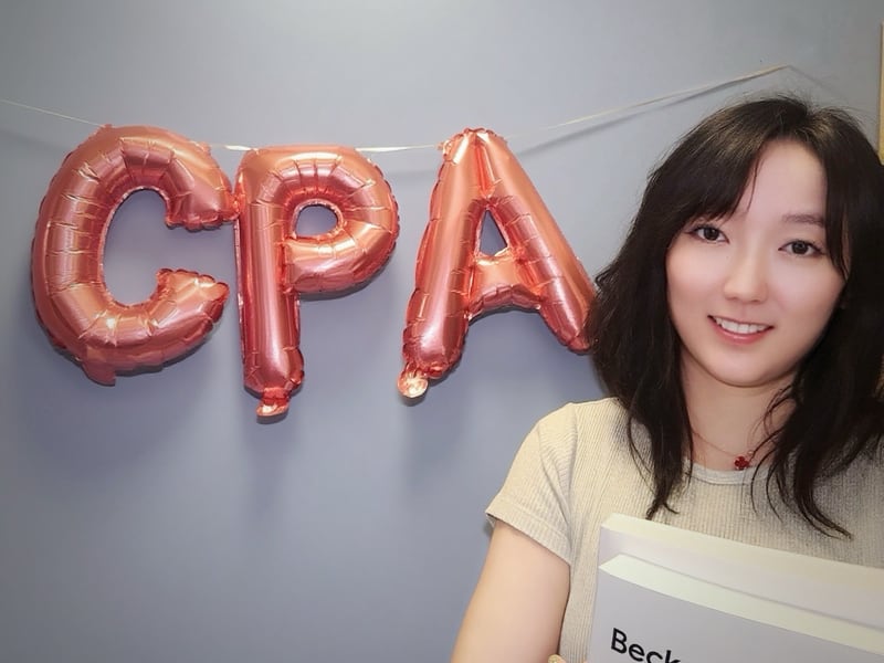 卢同学半身合影：抱 Becker USCPA 教材，背景粉色 CPA 气球字