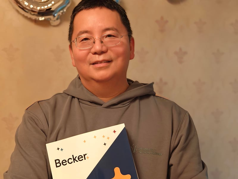马同学家中合影：戴眼镜手持 Becker FAR Financial 教材，背景银色 CPA 气球字